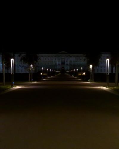 A night-time view of a long, straight, illuminated avenue lined with modern streetlights leading directly toward the brightly lit facade of a massive neoclassical palace.