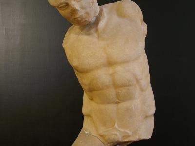 A marble torso of a male warrior statue, displayed on a metal stand against a dark background, with the head missing and one leg broken off at the thigh.