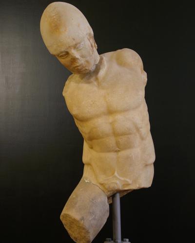 A marble torso of a male warrior statue, displayed on a metal stand against a dark background, with the head missing and one leg broken off at the thigh.