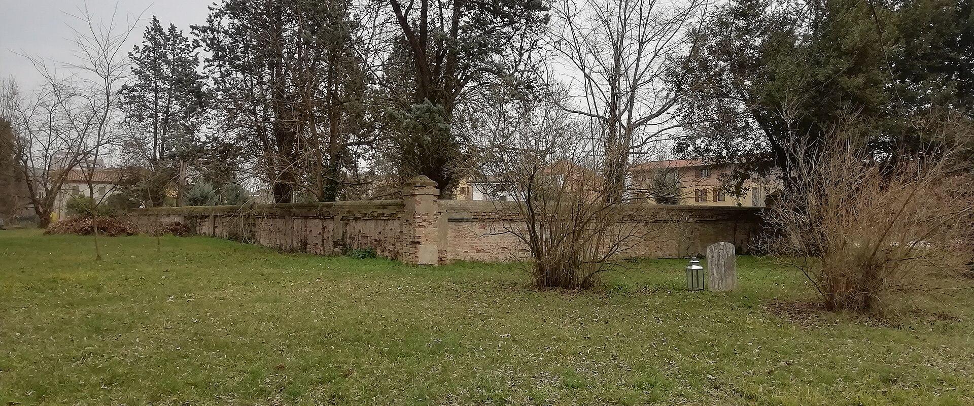 A grassy outdoor area enclosed by a low brick wall, with trees, a single standing stone marker, and a lantern on the ground; no visible signage or identifying text.