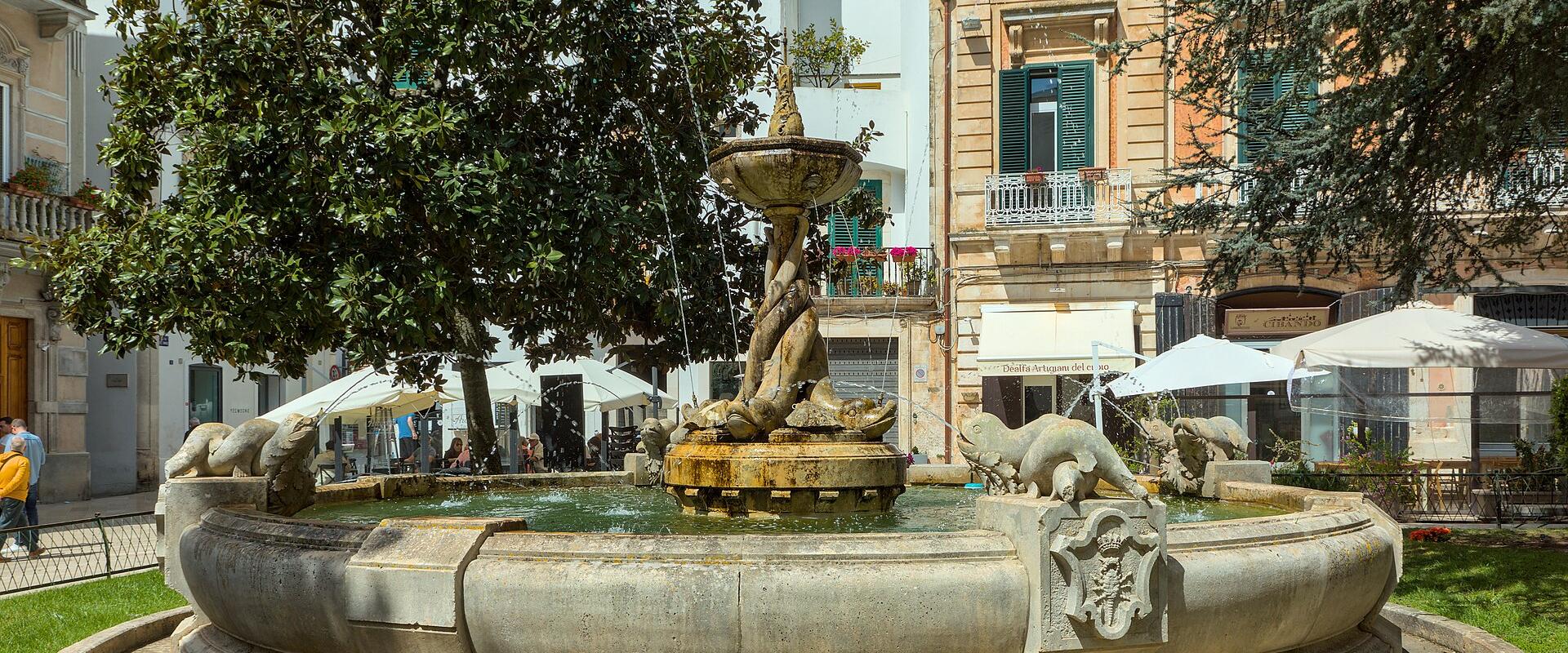 A large, ornate stone fountain with sculpted lions and a central twisted column, situated in a grassy public square surrounded by multi-story buildings with balconies and outdoor café umbrellas.