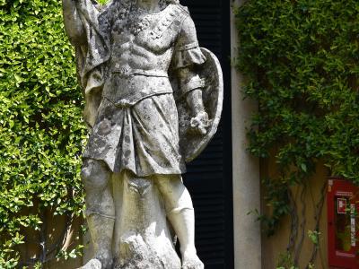 A weathered stone statue of a Roman soldier holding a shield and sword, standing on a pedestal in a garden with yellow poppies and ivy-covered walls.