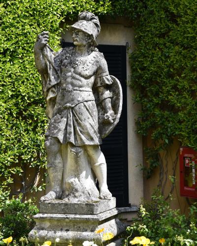 A weathered stone statue of a Roman soldier holding a shield and sword, standing on a pedestal in a garden with yellow poppies and ivy-covered walls.