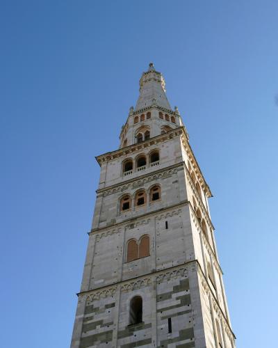 A low-angle photograph of a tall, ornate stone bell tower with multiple tiers of arched windows and decorative blind arcades, set against a clear blue sky.