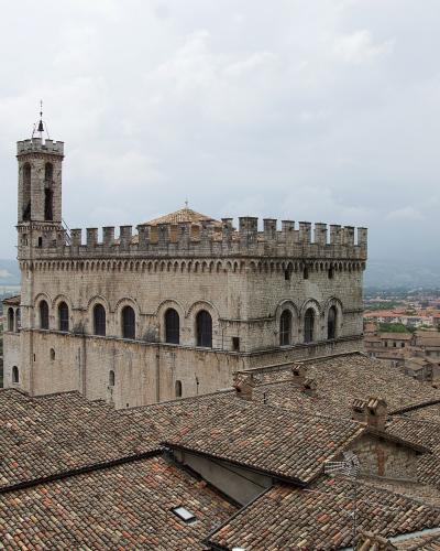 A high-angle view of a large, medieval stone palace with a prominent tower and crenellated roofline, set against a backdrop of terracotta-tiled rooftops and distant hills under an overcast sky.