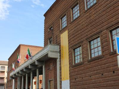 A low-angle exterior shot of a large, modern red-brick building with a colonnaded entrance portico, Italian flags, and a yellow vertical banner reading "STRADIVARI festival".