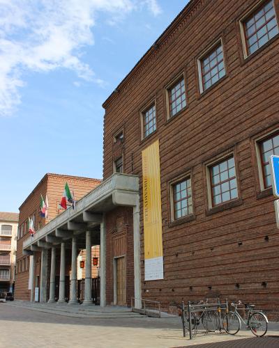 A low-angle exterior shot of a large, modern red-brick building with a colonnaded entrance portico, Italian flags, and a yellow vertical banner reading "STRADIVARI festival".