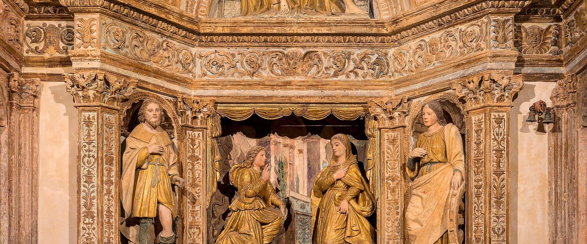 An ornate Baroque-style altar with gilded statues, including a Pietà above and figures below, set within a richly decorated stone archway featuring intricate carvings and a coffered ceiling.