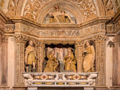 An ornate Baroque-style altar with gilded statues, including a Pietà above and figures below, set within a richly decorated stone archway featuring intricate carvings and a coffered ceiling.