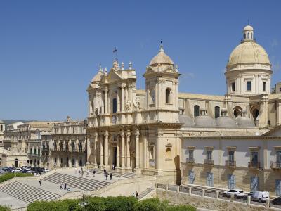 A wide-angle, color photograph of a large, ornate cathedral with a prominent central dome and twin bell towers, situated atop a grand staircase in a sunlit Italian town square.