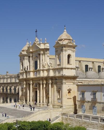 A wide-angle, color photograph of a large, ornate cathedral with a prominent central dome and twin bell towers, situated atop a grand staircase in a sunlit Italian town square.