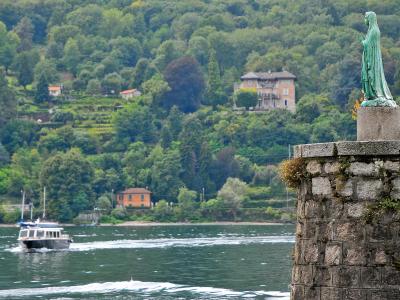 A view of a lake with a passenger boat moving across the water, a stone pier with a green statue of a robed figure in the foreground, and a lush, terraced hillside with villas in the background.