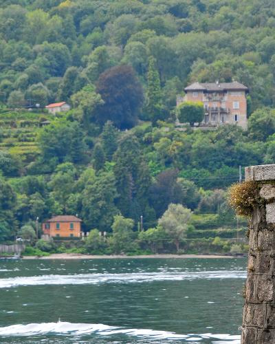 A view of a lake with a passenger boat moving across the water, a stone pier with a green statue of a robed figure in the foreground, and a lush, terraced hillside with villas in the background.