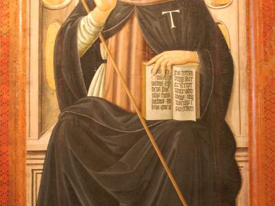 A framed Renaissance-style painting depicting a seated, bearded male saint in black monastic robes holding a staff and an open book, with a small black dog at his feet and a bell hanging in the background.