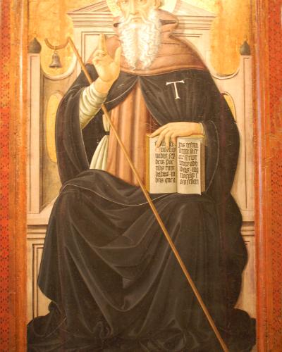 A framed Renaissance-style painting depicting a seated, bearded male saint in black monastic robes holding a staff and an open book, with a small black dog at his feet and a bell hanging in the background.