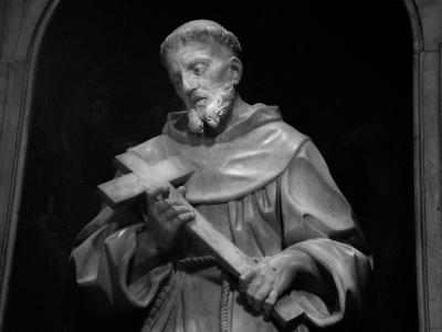 A black-and-white photograph of a stone statue depicting a bearded male saint in Franciscan robes holding a large cross, set within a dark arched niche.