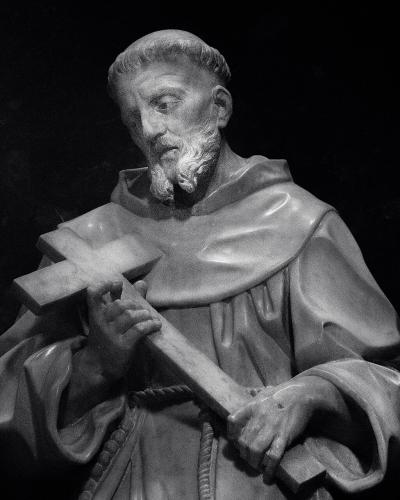 A black-and-white photograph of a stone statue depicting a bearded male saint in Franciscan robes holding a large cross, set within a dark arched niche.