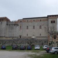 A large medieval stone fortress with crenellated towers and arched windows, situated on a raised stone platform with a gravel parking lot in front containing several modern cars; tall cypress trees are visible to the left, and a smaller brick stru