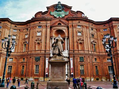 The image shows the exterior of a large, ornate red-brick palace with a prominent statue in the foreground and a plaque reading "VITTORIO EMANUELE II" above the central archway.