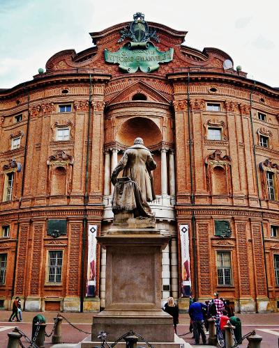 The image shows the exterior of a large, ornate red-brick palace with a prominent statue in the foreground and a plaque reading "VITTORIO EMANUELE II" above the central archway.