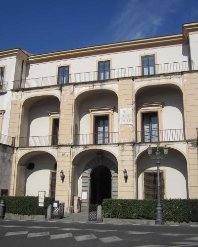 A large, multi-story historic building with arched ground-floor loggia, balconies, and a plaque reading "MUSEO CORREALE DI TERRANOVA" above the central entrance, set against a clear blue sky with street-level hedges and a lamppost in front.