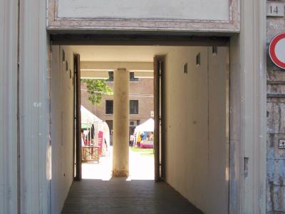 A view through a large, arched stone gateway into a courtyard where market stalls are set up under white tents, with a prominent stone column visible in the center of the passage.