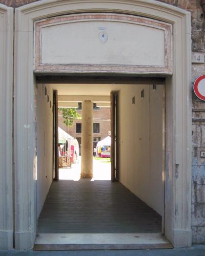 A view through a large, arched stone gateway into a courtyard where market stalls are set up under white tents, with a prominent stone column visible in the center of the passage.