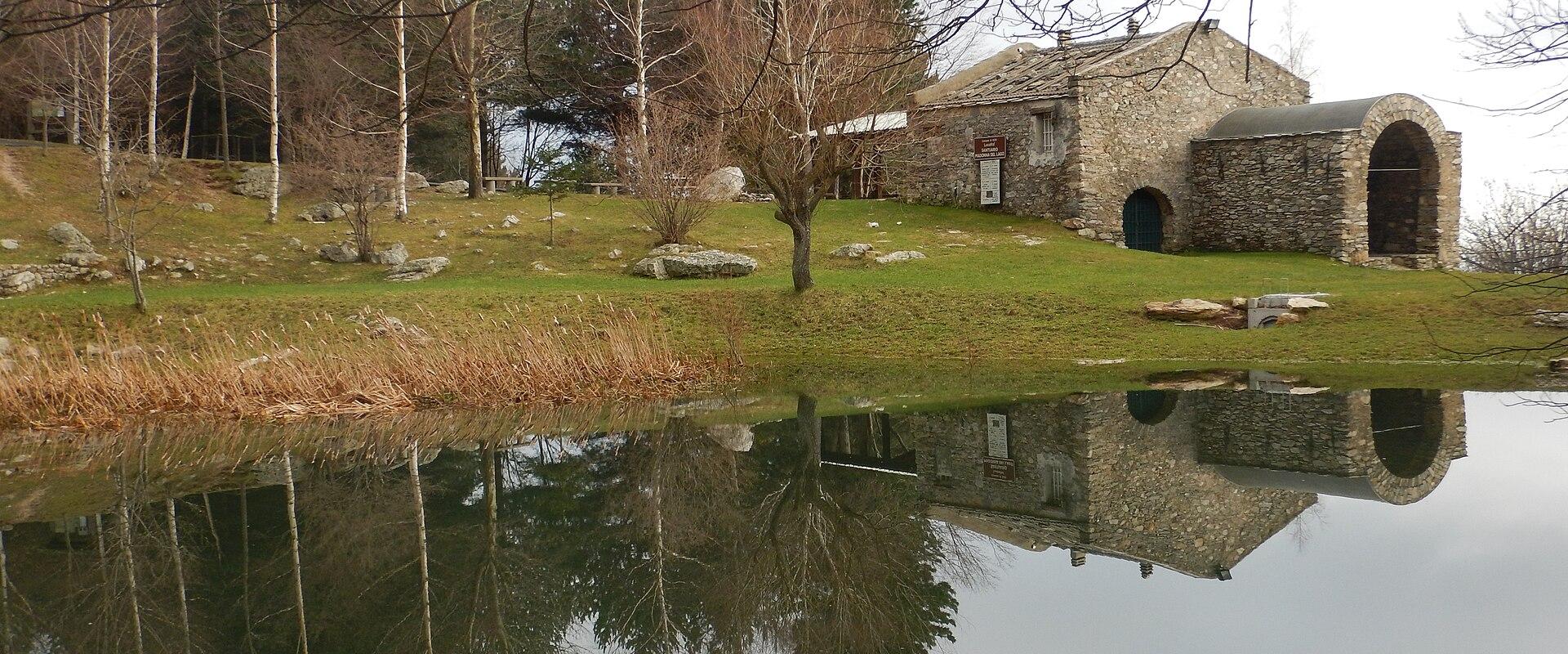 A stone building with arched openings sits beside a calm body of water, its reflection mirrored perfectly; bare trees and grassy slopes surround the scene under an overcast sky.