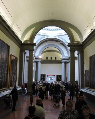 A long, crowded gallery hall with large framed paintings on the walls, arched ceilings, and visitors walking through; no visible signage or specific artwork labels are legible.