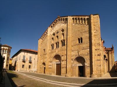 A low-angle photograph of a large, ancient stone basilica with Lombard Romanesque features including blind arcades, sculpted portals, and a prominent cross on the facade, set against a clear blue sky in an urban plaza.
