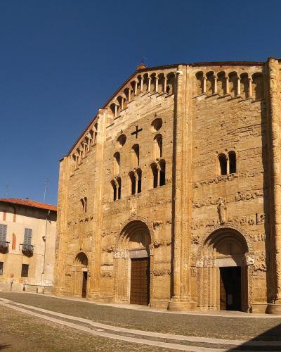 A low-angle photograph of a large, ancient stone basilica with Lombard Romanesque features including blind arcades, sculpted portals, and a prominent cross on the facade, set against a clear blue sky in an urban plaza.