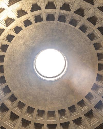 A low-angle photograph looking directly up into the coffered interior of a massive concrete dome, centered on the circular oculus opening to the sky.