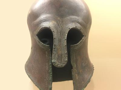 A bronze Corinthian-style helmet displayed on a stand against a plain, light-colored background, with visible decorative engravings around the eye and cheek openings.