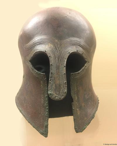 A bronze Corinthian-style helmet displayed on a stand against a plain, light-colored background, with visible decorative engravings around the eye and cheek openings.