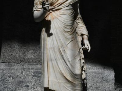 A full-length marble statue of a draped female figure, likely from antiquity, displayed on a pedestal against a rough stone wall in a dimly lit indoor setting.
