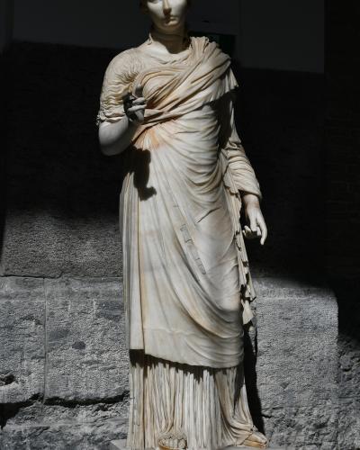 A full-length marble statue of a draped female figure, likely from antiquity, displayed on a pedestal against a rough stone wall in a dimly lit indoor setting.