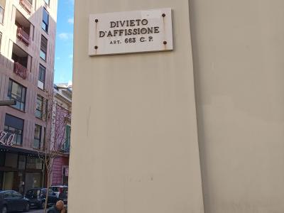 A close-up of a street sign mounted on a beige wall, clearly reading "VIA SPARANO DA BARI" with a municipal crest, alongside a "DIVIETO D'AFFISSIONE" notice, with a street scene visible in the background.