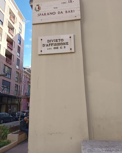 A close-up of a street sign mounted on a beige wall, clearly reading "VIA SPARANO DA BARI" with a municipal crest, alongside a "DIVIETO D'AFFISSIONE" notice, with a street scene visible in the background.