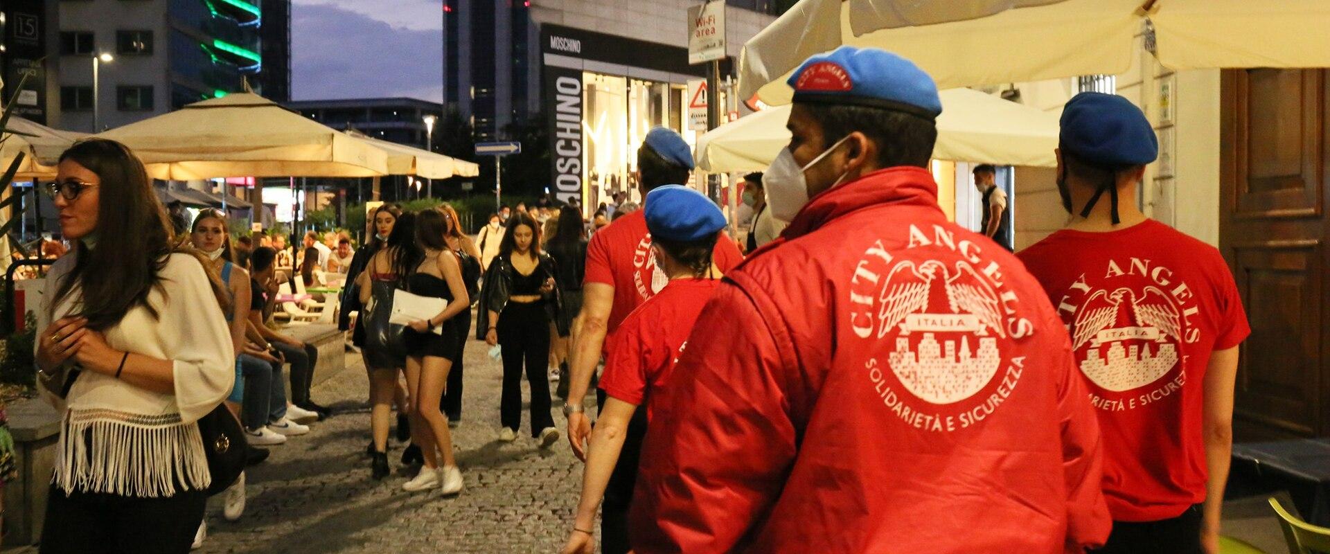 A group of security personnel wearing red jackets and blue berets with "CITY ANGELS" and "SOLIDARIETÀ E SICUREZZA" on the back, walking through a cobblestone street lined with outdoor cafe umbrellas and pedestrians at dusk.