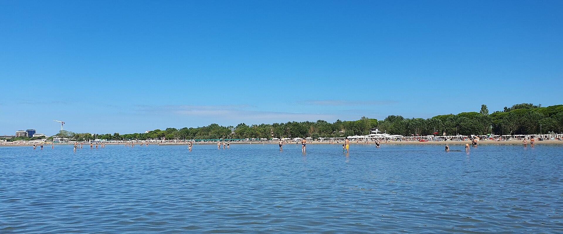 A wide, calm body of water with people wading and swimming near a distant shoreline lined with trees, beach umbrellas, and buildings under a clear blue sky.