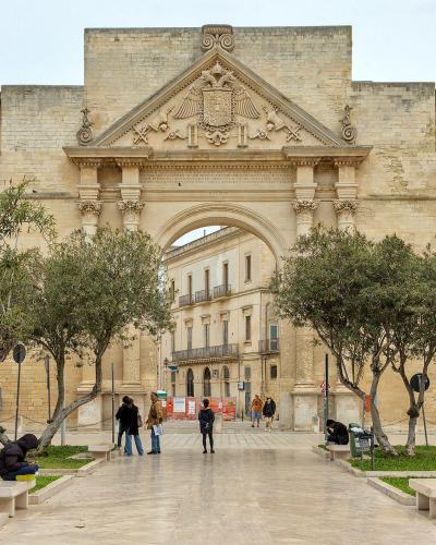 A large, ornate stone triumphal arch with a central passageway, flanked by trees and benches in a paved public square, featuring a coat of arms and inscriptions on its facade.