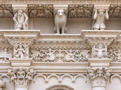 A close-up architectural detail of an ornate stone facade featuring sculpted figures including a lion, atlantes, cherubs, and mythological creatures in high relief, characteristic of Baroque stonework.