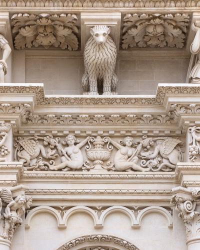 A close-up architectural detail of an ornate stone facade featuring sculpted figures including a lion, atlantes, cherubs, and mythological creatures in high relief, characteristic of Baroque stonework.