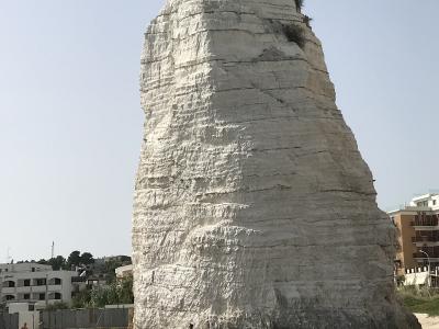 A tall, white, conical monolith of layered rock rising from a sandy beach with people at its base and coastal buildings in the background.