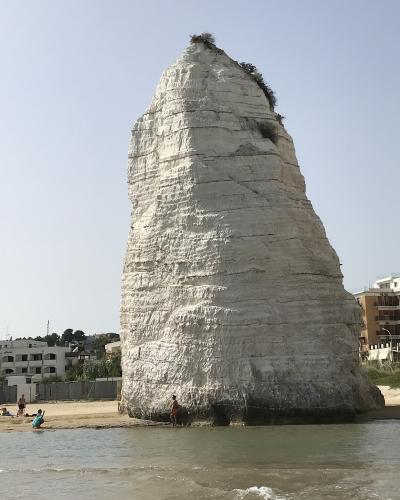 A tall, white, conical monolith of layered rock rising from a sandy beach with people at its base and coastal buildings in the background.