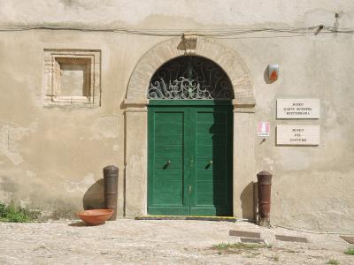The image shows the entrance to a building with a green double door under an arched brick frame, flanked by two old cannons and a terracotta bowl, with stone plaques on the wall reading "MUSEO D'ARTE MODERNA 'MEDITERRANIA'" and "MUSEO DEL COSTUME"