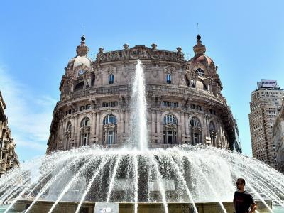 A low-angle shot of a large, ornate fountain with a central jet and wide spray, set in a city square surrounded by grand, historic buildings including one with "BANCA" visible on its facade and another with a dome.
