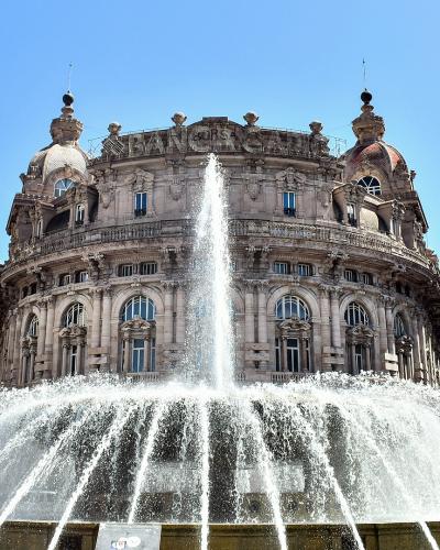 A low-angle shot of a large, ornate fountain with a central jet and wide spray, set in a city square surrounded by grand, historic buildings including one with "BANCA" visible on its facade and another with a dome.