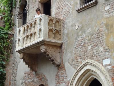 A low-angle view of a weathered brick and plaster building facade featuring a distinctive stone balcony with Gothic-style arches, ivy climbing the wall to the left, and a woman standing on the balcony.
