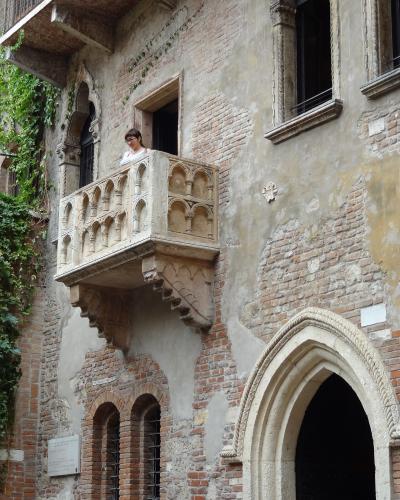 A low-angle view of a weathered brick and plaster building facade featuring a distinctive stone balcony with Gothic-style arches, ivy climbing the wall to the left, and a woman standing on the balcony.